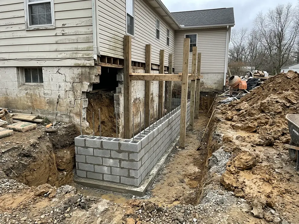 Excavated trench beside an older house with a section of new concrete block wall laid on the footing and wood shoring posts